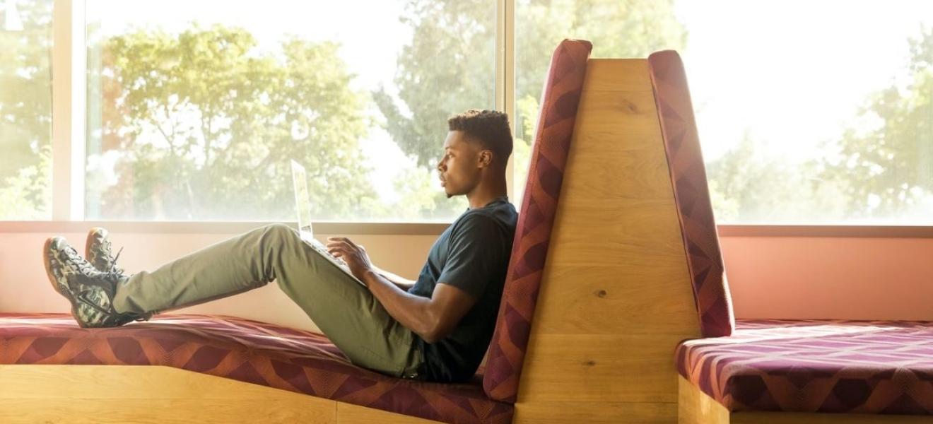 A man sitting on a bench seat using a laptop. Behind him is a window through which trees can be seen