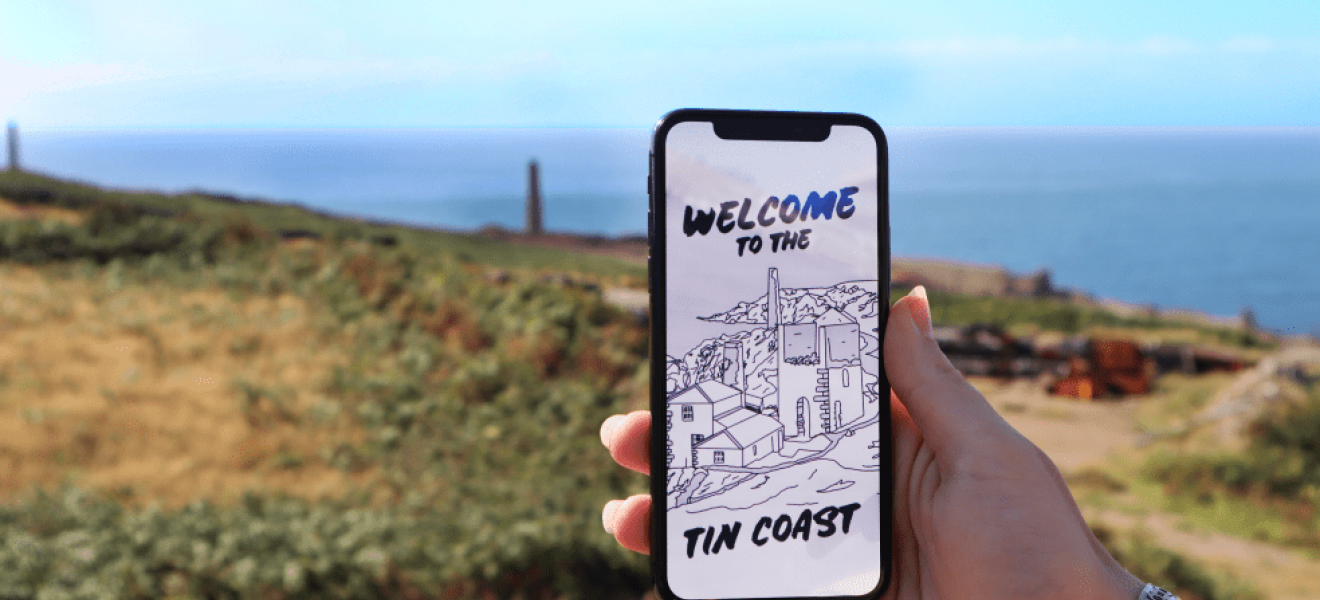 Explore more of the tin coast using the XplorTINCOAST app A hand holds a phone showing the XplorTINCOAST landing page. There is a coastal background.