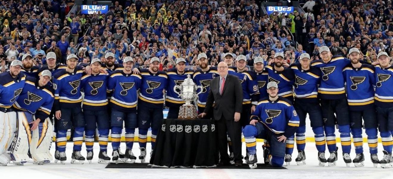 the St Louis blues team in front of the NHL cup on the ice, with their fans behind them