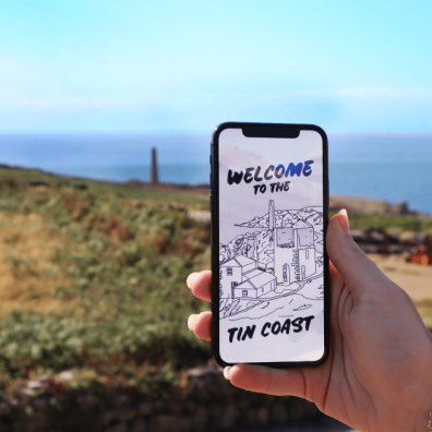 A hand holds a phone showing the XplorTINCOAST landing page. There is a coastal background.