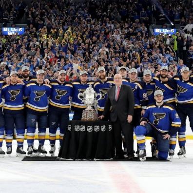 the St Louis blues team in front of the NHL cup on the ice, with their fans behind them